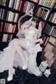 A woman in a wedding dress sitting on a chair in front of a bookcase.